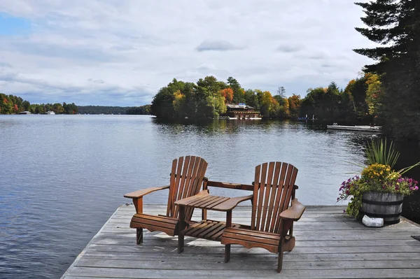 İki Muskoka sandalyesi sakin bir göle bakan ahşap bir rıhtımda oturuyor. Suyun karşısında yeşil ağaçların arasına kurulmuş bir kulübe var..