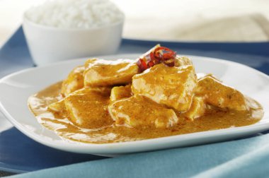 Beyaz bir tabakta sıcak köri soslu doğranmış tavuk ile mavi bir sırt grubunda bir kase pirinç servis edilir.