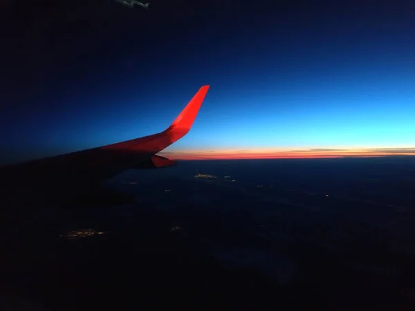 The bright red wing tip of an airplane against the dark evening sky with a red sunset on the horizon.