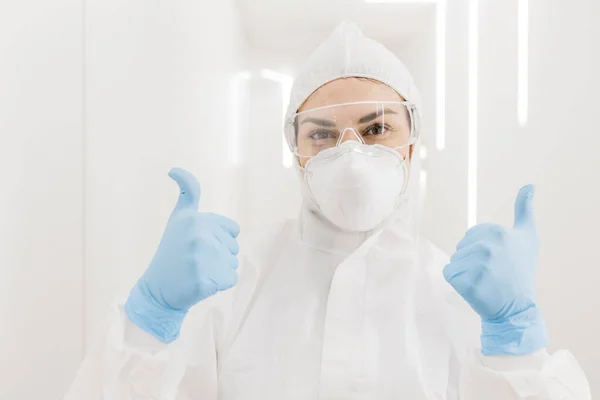 Kciuki w niebieskich rękawiczkach medycznych. Dziewczęcy lekarz w kombinezonie ochronnym podczas epidemii w białym korytarzu nowoczesnego szpitala. Zabawa. — Zdjęcie stockowe