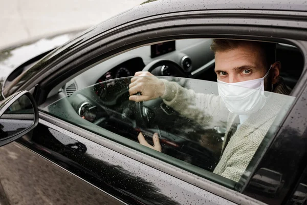 Mężczyzna w masce medycznej w samochodzie patrzący w kamerę. Kierowca podczas epidemii. Ochrona przed wirusem. Kierowca w kurtce w samochodzie. Koronawirus, choroba, zakażenie, kwarantanna, kovid-19 — Zdjęcie stockowe