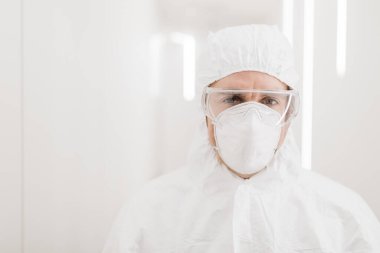 Hastanenin beyaz modern koridorunda salgın sırasında koruyucu kıyafetli bir doktorun erkek portresi..