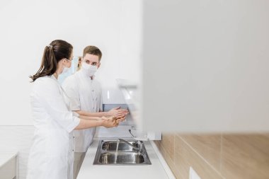 İki doktor ellerini modern beyaz bir dolapta salgın sırasında koruyucu tıbbi maskelerle yıkıyor.. 