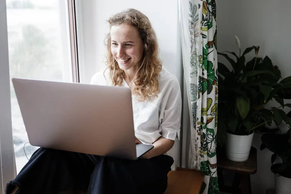 漂亮的女孩坐在一个窗户上，在一个现代化的公寓里带着笔记本电脑，通过视频链接进行交流 图库照片