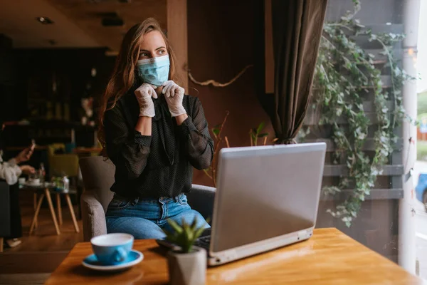 Maskeli ve eldivenli güzel bir beyaz iş kadını bir kafede oturuyor, kahve içiyor ve bir dizüstü bilgisayar üzerinde çalışıyor. COVID - 19 virüs koruması