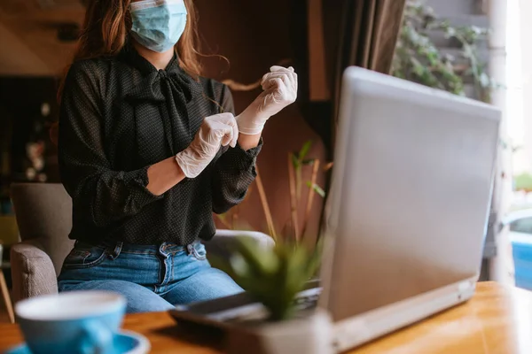 A beautiful caucasian business woman with a mask and gloves is sitting in a cafe, drinking coffee and working on a laptop. COVID - 19 virus protection at work