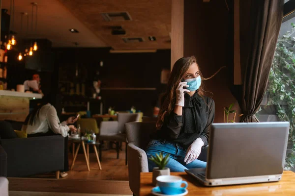 Maskeli ve eldivenli güzel beyaz bir kadın bir kafede oturuyor ve telefonu kullanıyor. COVID virüs koruması - 19