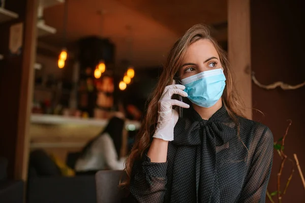 Maskeli ve eldivenli güzel beyaz bir kadın bir kafede oturuyor ve telefonu kullanıyor. COVID virüs koruması - 19