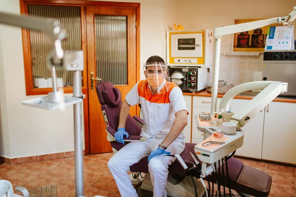 Diş muayenehanesinde maske ve güneşlik takan beyaz bir dişçinin portresi. COVID - 19 virüs koruması