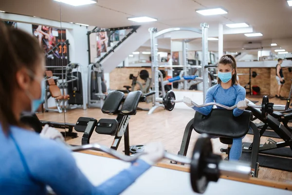 Spor salonunda ağırlıkları olan koruyucu maskeli beyaz bir kadın.