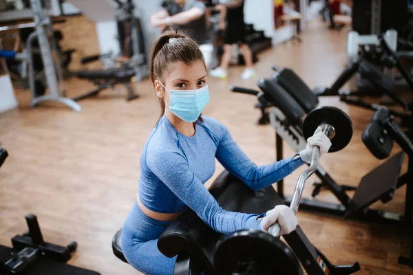 Spor salonunda ağırlıkları olan koruyucu maskeli beyaz bir kadın.