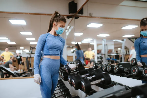Spor salonunda ağırlıkları olan koruyucu maskeli beyaz bir kadın.