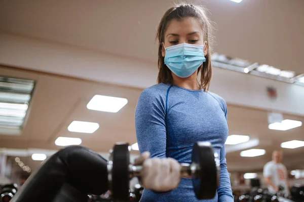 Spor salonunda ağırlıkları olan koruyucu maskeli beyaz bir kadın.