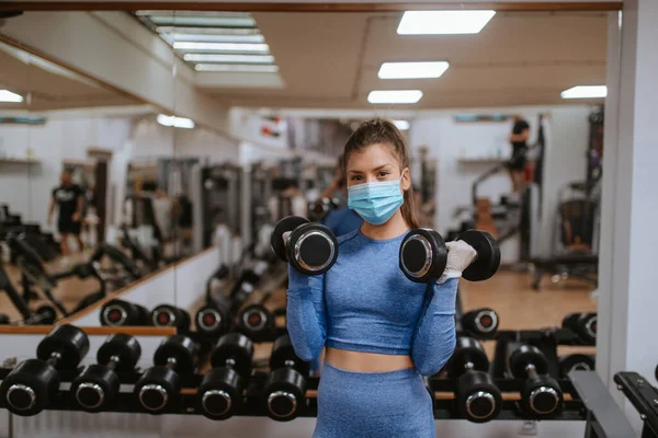Spor salonunda ağırlıkları olan koruyucu maskeli beyaz bir kadın.