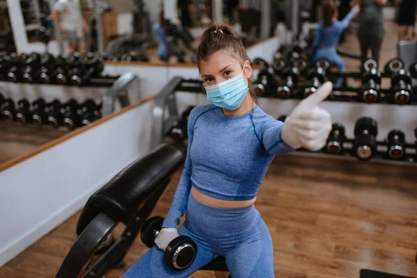 Spor salonunda ağırlıkları olan koruyucu maskeli beyaz bir kadın.