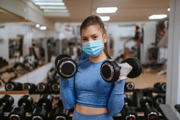 Spor salonunda ağırlıkları olan koruyucu maskeli beyaz bir kadın.