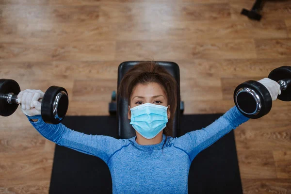 Spor salonunda ağırlıkları olan koruyucu maskeli beyaz bir kadın.