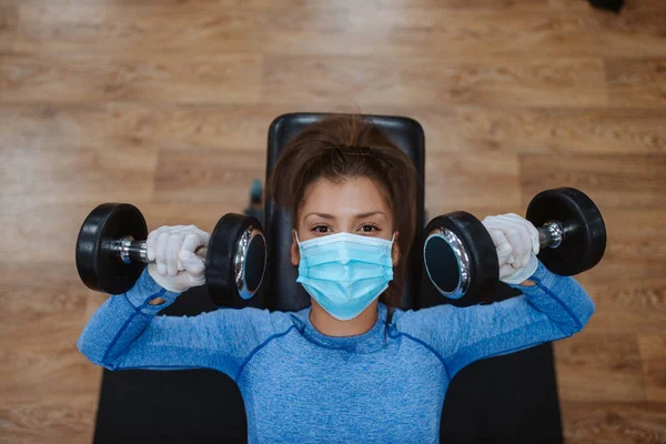 Spor salonunda ağırlıkları olan koruyucu maskeli beyaz bir kadın.