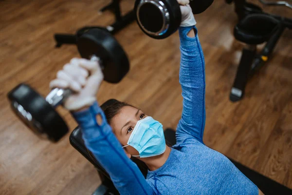 Spor salonunda ağırlıkları olan koruyucu maskeli beyaz bir kadın.
