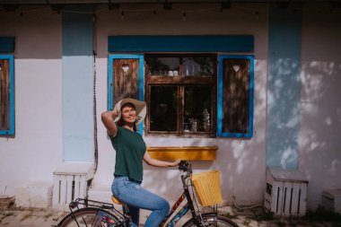 Hasır şapkalı güzel, beyaz bir kadın elinde sepetle bir kır evinin yanında bisiklet sürüyor.