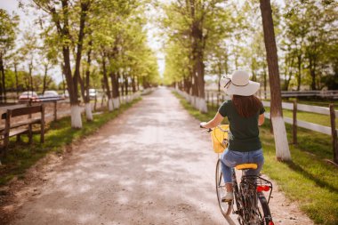 Hasır şapkalı güzel beyaz bir kadının arka tarafı kırsalda sepeti olan bir bisiklete biniyor.