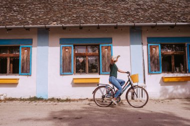 Hasır şapkalı güzel, beyaz bir kadın elinde sepetle bir kır evinin yanında bisiklet sürüyor.