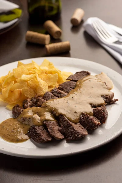 Steak with sauce with potato chips on white plate and wine glass, cork, silverware and napkin