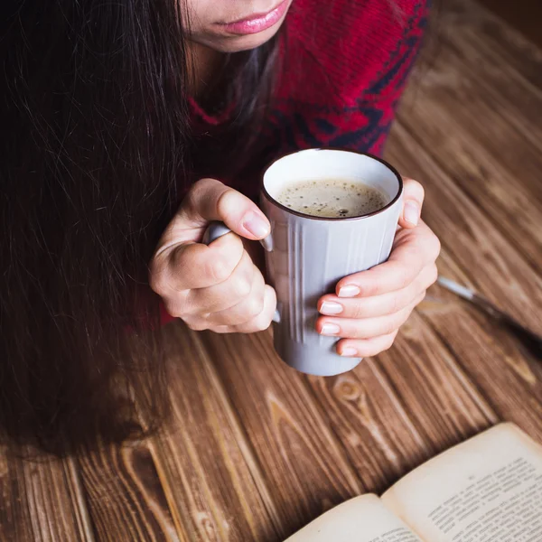 Молодая женщина в красном свитере держит чашку кофе и читает книгу — стоковое фото