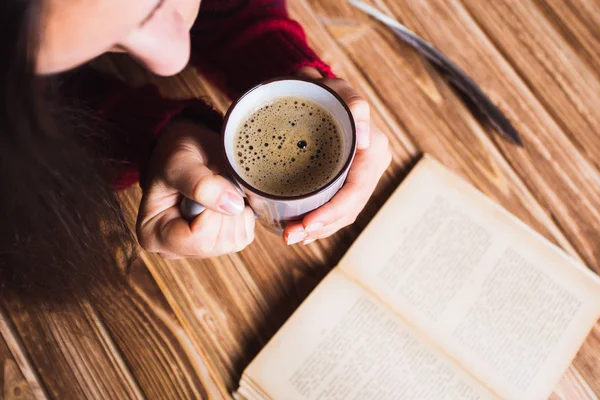 Молодая женщина в красном свитере держит чашку кофе и читает книгу — стоковое фото