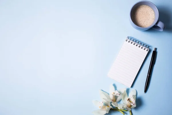 Tasse de café et carnet avec stylo et fleur d'orchidée sur fond bleu, vue de dessus — Photo