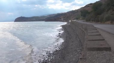 Deniz ve dağlar arasında dar bir yol var. Dalgalar kayalık sahile vurdu. Yol, dalgakıran tarafından sudan ayrılır. Ağaçlar kuru tepelerde yetişir. Gökyüzü bulutların arkasında gizleniyor. Çatı rafı olan koyu renk araba geçiyor.