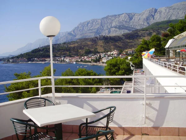 Otelin manzarası. Ön planda kahve sehpası olan sandalyeler görebiliyoruz, sağ tarafında otel yüzme havuzu olan sandalyeler. Bir manzara çam ağaçlarının üzerinden mavi denize ve arka planda pervazları ve dağları olan bir körfeze götürür..