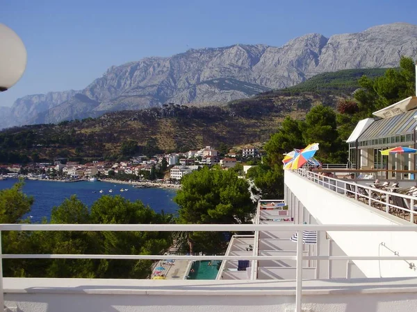 Çam ağaçlarının üzerinden bir manzara. Tepenin altında küçük hayalet evleri, bir tarafında kaya, diğer tarafında yüzme havuzu ve renkli şemsiyeleri olan otel terasları olan körfezi görüyoruz..