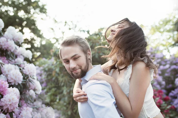 Emotional Love story of a young couple. Woman is sitting on a man\'s shoulders in a rhododendrons park in England behind pink blooming bush, tree. Woman is wearing white top, man is wearing light blue shirt.