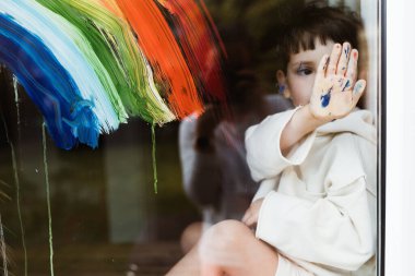 Küçük çocuk karantina sırasında pencereye gökkuşağı çiziyor ve pencereden dışarı bakıyor. Kirli eller. Hepimiz iyi olalım..