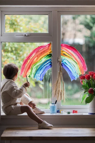 Little boy painting rainbow on a window and looking through the window while sitting home during quarantine.  Let\'s all be well. Quarantine. Virus. Pandemic.Coronavirus. Stay home concept. Support NHS. Thank you NHS.