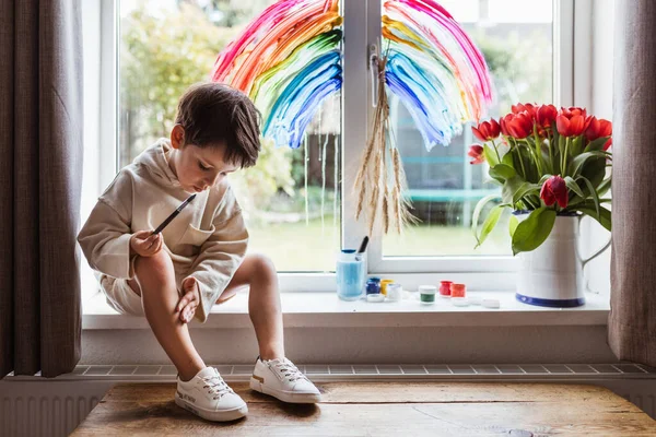 Little boy painting rainbow on a window while sitting home during quarantine. Dirty hands. Let\'s all be well. Quarantine. Virus. Pandemic.Coronavirus. Stay home concept. Support NHS. Thank you NHS