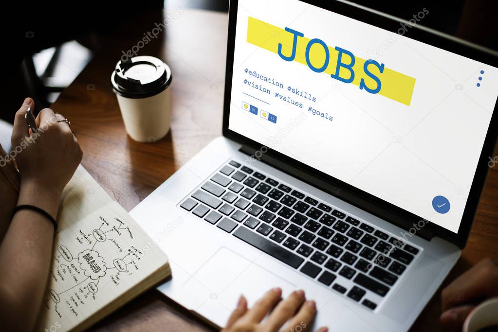 Wooden table with laptop, coffee mug and diary, people working and writing notes, monitor screen with title: Jobs
