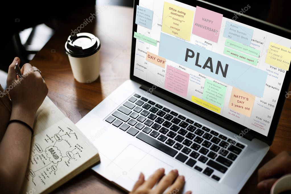 People working with laptop at table, person writing notes in diary, coffee mug on table, text on screen: Plan