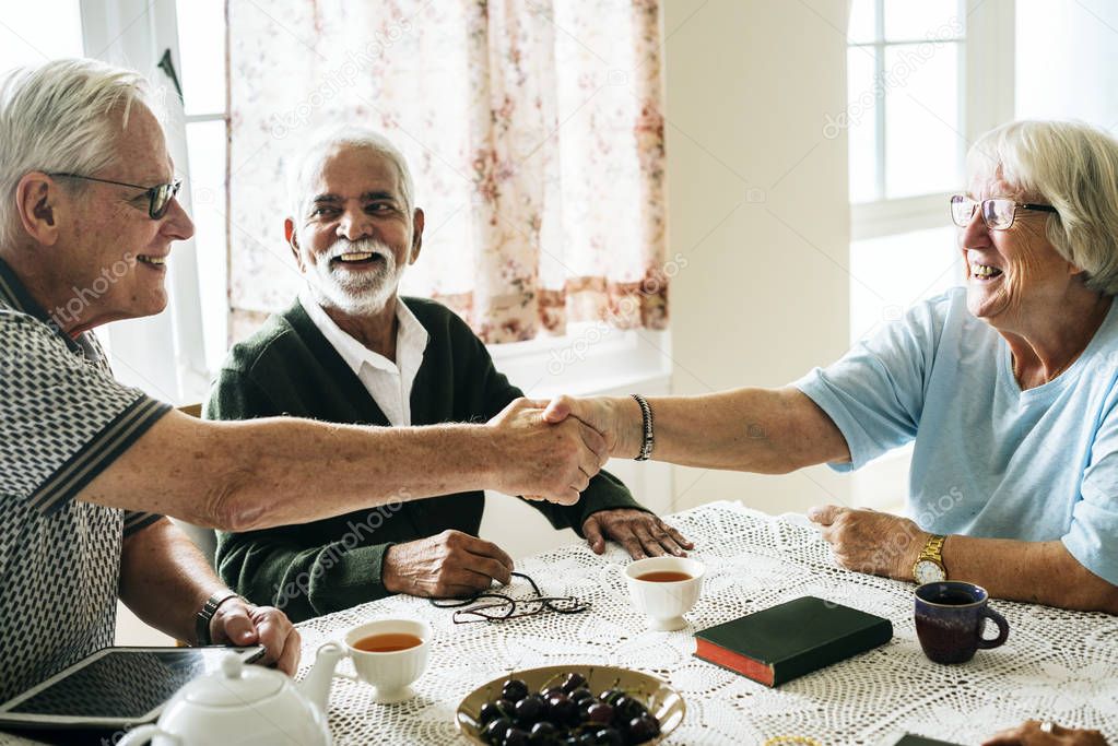 senior people shaking hands, sitting at table with tea at home 