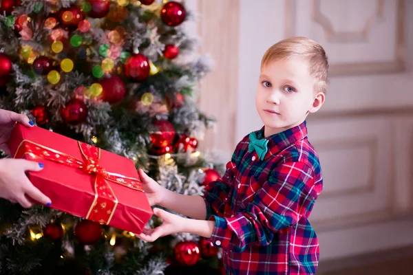 Woman give a cute boy in checkered shirt Christmas red gift box with Christmas tree on the background — Stock Photo, Image
