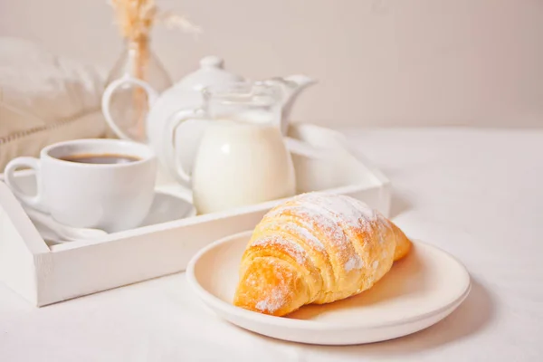 Fresh croissant bun on the white plate with cup of coffee, jar of milk on the wooden tray on the background. — Stock Photo, Image