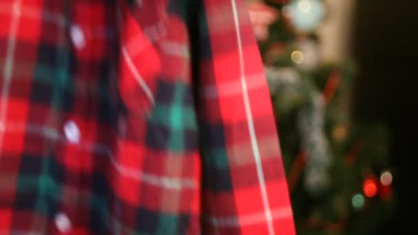 Mujer de la mano sostiene y se ve camisa a cuadros para el hijo para una fiesta de Navidad. Árbol de Navidad en el fondo . — Vídeos de Stock