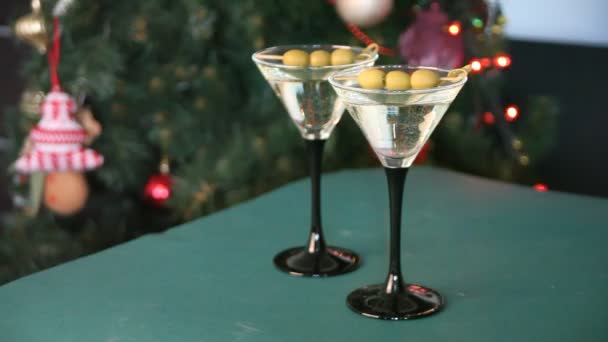 緑のテーブルの上にオリーブとギフトボックスとマティーニの2つのグラス 背景にはボケが入った軽いガーランドとクリスマスツリー 女性の手は3つの様々なギフトボックスを置く — ストック動画