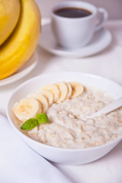 Овсяная каша с бананом в белой миске, чашка кофе на белом столе. Здоровье завтрак . — стоковое фото