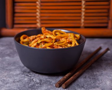 Udon eriştesi ve kase içinde tavuk eti. Koyu taş masada çubuklar var.