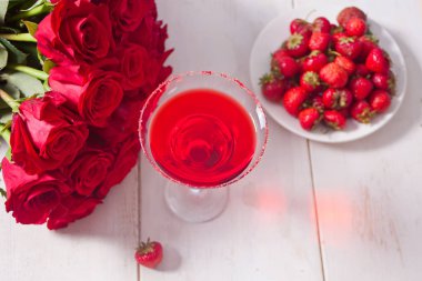 Temiz bardakta kırmızı egzotik alkollü kokteyl, taze çilekli tabak ve romantik yemek için ahşap beyaz masada kırmızı güller..