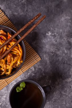 Udon eriştesi, kasede tavuk eti, yemek çubukları ve koyu taştan arka planda bir fincan nane çayı. Üst görünüm