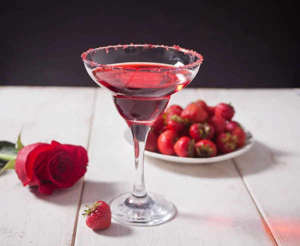 Красный экзотический алкогольный коктейль в прозрачном стакане, тарелка со свежей клубникой и красная роза на деревянном белом столе для романтического ужина . — стоковое фото