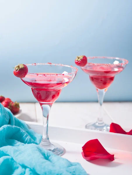 Czerwony egzotyczny koktajl alkoholowy w przezroczystych okularach i czerwone płatki róż na drewnianym białym stole na romantyczną kolację. — Zdjęcie stockowe